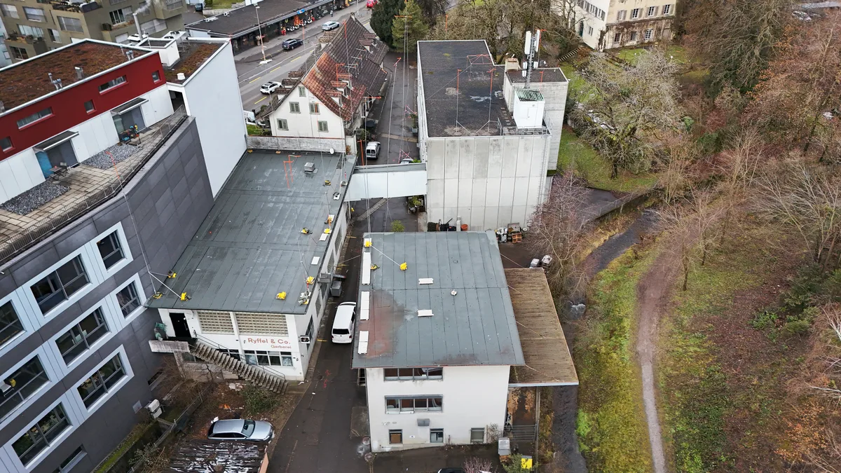 Die Fläche des Areals ist kleiner, als dies für eine Arealüberbauung nötig wäre. Rechts sieht man den Schlossbach. Drohnenaufnahme eines Areals mit Gebäuden und einem Bach.